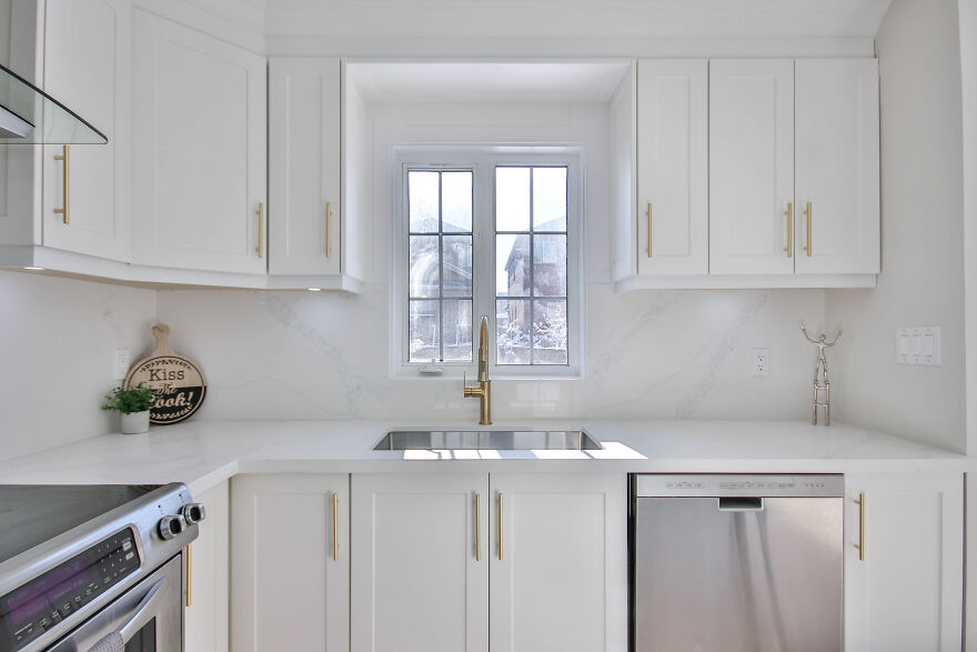 White modern kitchen cabinets with gold handles and a stainless steel dishwasher under bright natural light.