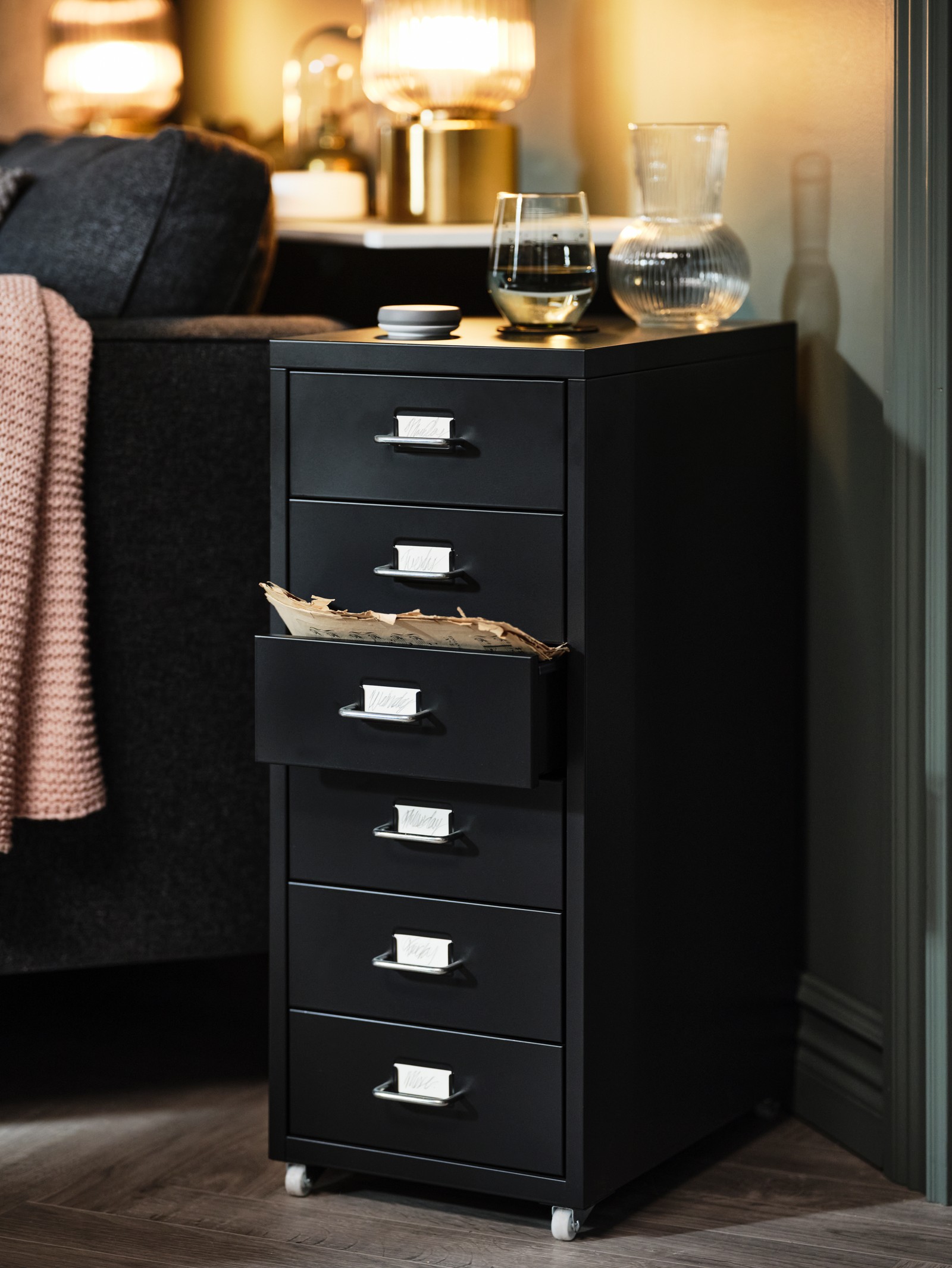 A black HELMER drawer unit on castors stands at an angle next to a sofa. Two lit brass lamps are on a shelf behind the s