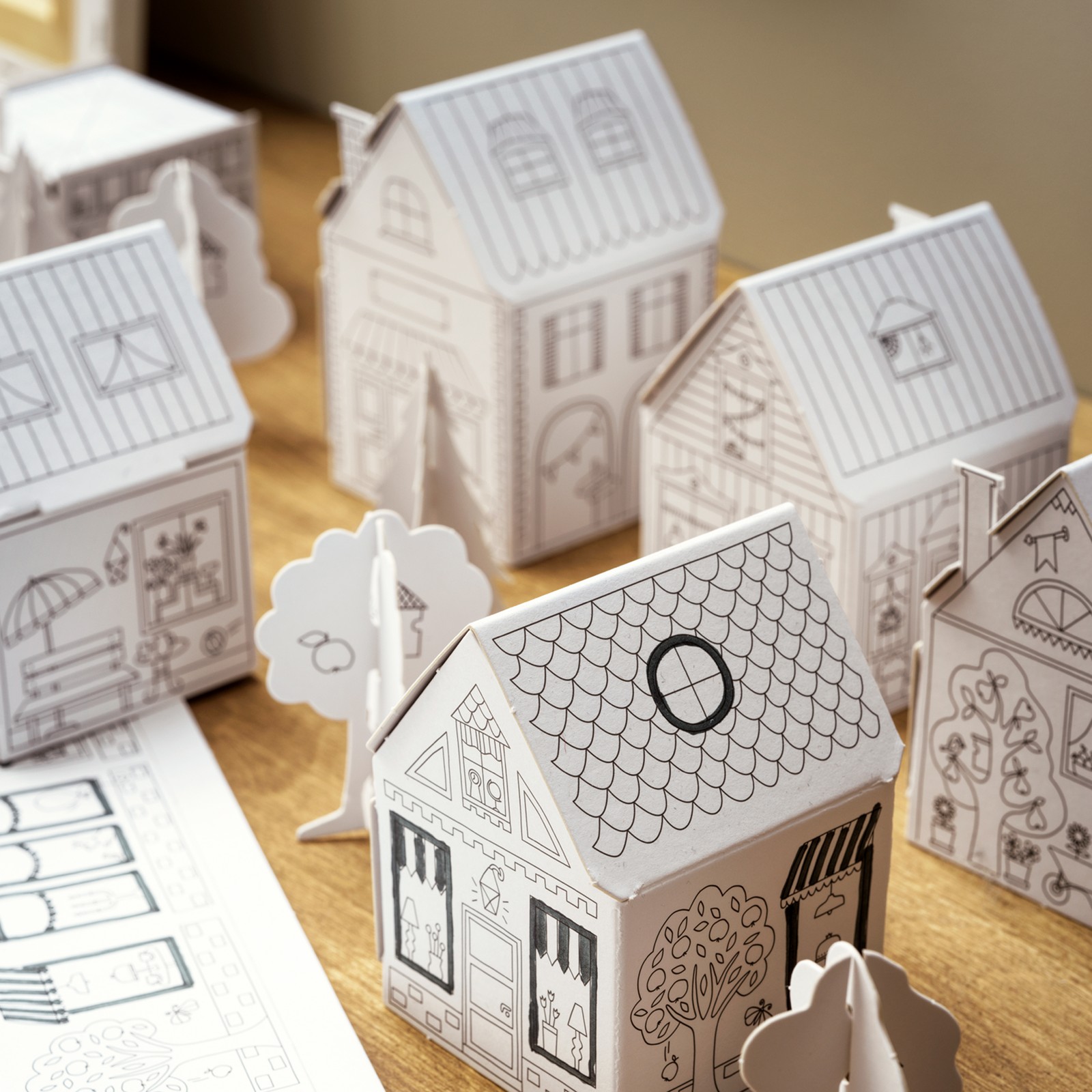 A children‘s toy MÅLA cardboard town with different houses and trees, placed on a wooden surface.