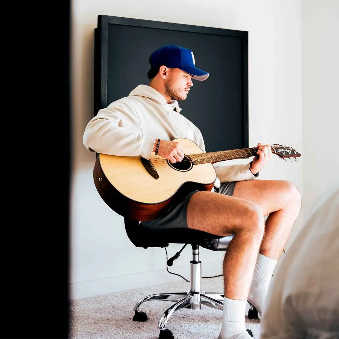 A young man is playing the guitar while sitting on his chair away from his black wall-mounted desk.